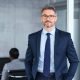 istockphoto-1413766112-612×612 Portrait of handsome mid adult business man standing in modern office. Successful mature entrepreneur in formal clothing looking at camera with satisfaction. Confident man in suit with eyeglasses and beard standing with hands in pocket and looking at camera in office.